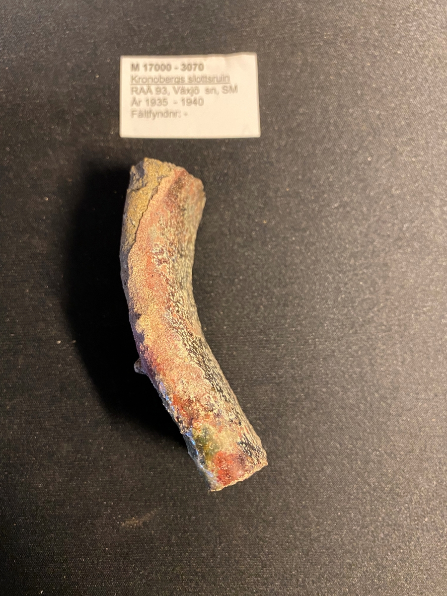 Fragment av en rak mynningsbit till en degel. Sekundärt bränd med delvis cintrade röda och gräna ytor på in- och utsidan. Relativt grovt gods. Liknar M 17000-3066.