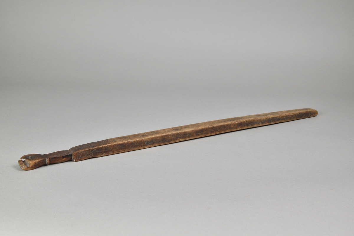 Träaln tillverkad i björk. Fyrsidig med rundat, nött avslut. Kort handtag, avslutat med snidad hand med hål för upphängning. Nedanför handtaget inskuret årtal, "1726".