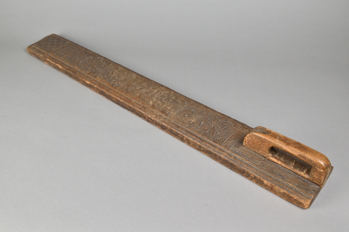 Mangelbräde tillverkat i trä. Rak med profilerade långsidor och handtag av trä. Ovansidan rikt ornerad med skurna mönster, samt inskriften "Anno 1770".