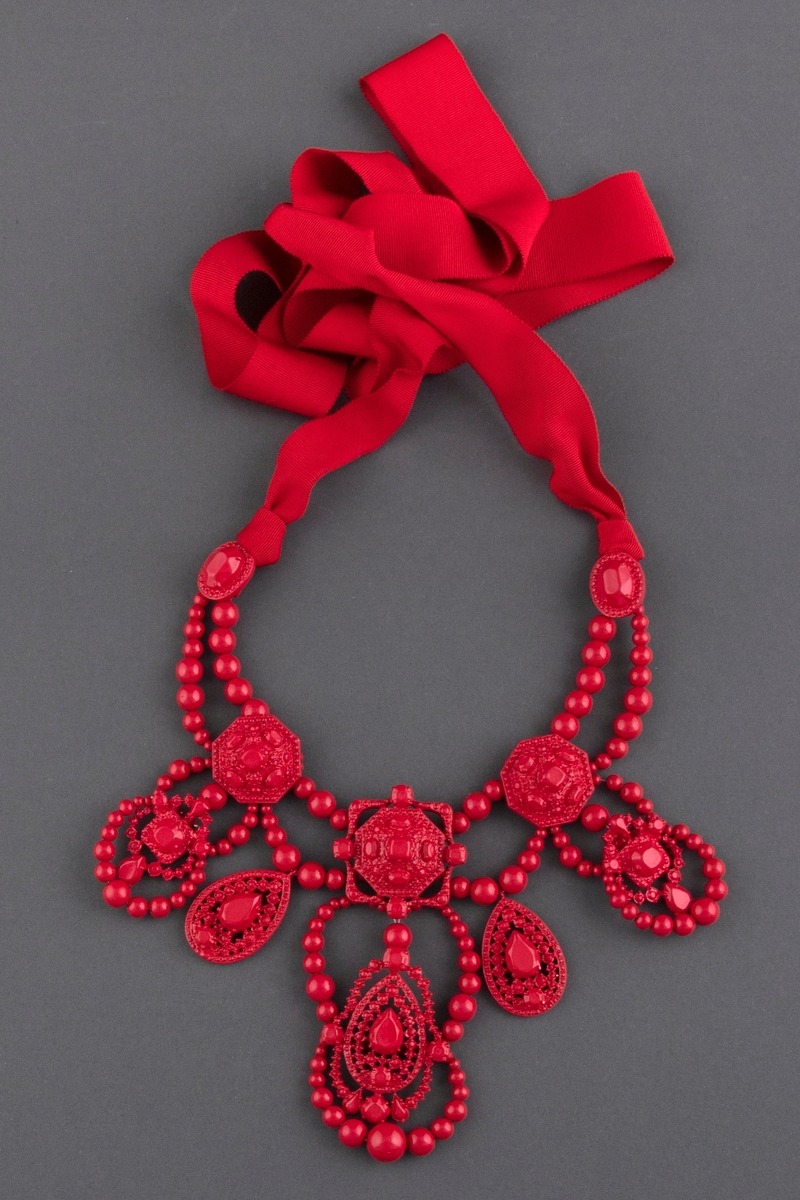 Stort halssmykke av rødlakkert metall, plastperler og brede tekstilbånd i samme farge. Smykket knytes i nakken.