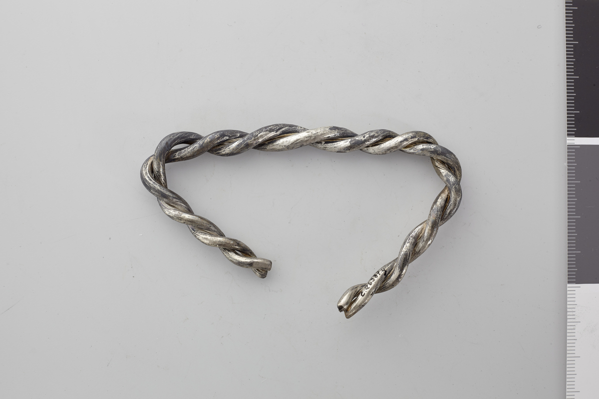 Forbøiet håndleddsring av sølv, dannet av 2 tykke sølvtener, som er avklippet i begge ender. Største nuværende tverrmål 9 cm, største tykkelse 0,9 cm. 