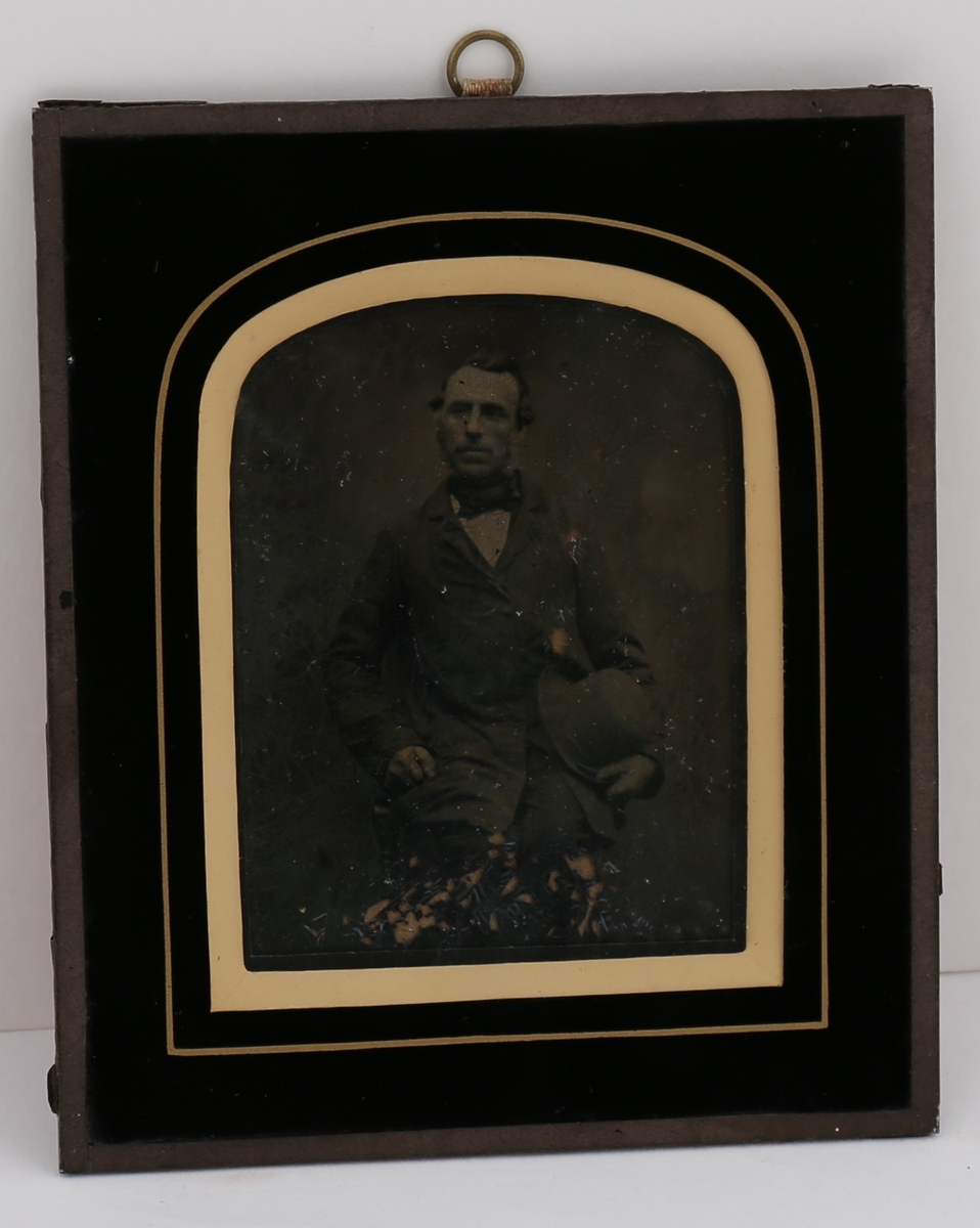 Portrett av mann i mørk dress med kalvekryss i halsen og hatten i hånden. Ambrotyp fototeknikk. Dette bildet er en glassplate der motivet er positivt fremkalt fra et glassplate-negativ. På baksiden en opphengsring.