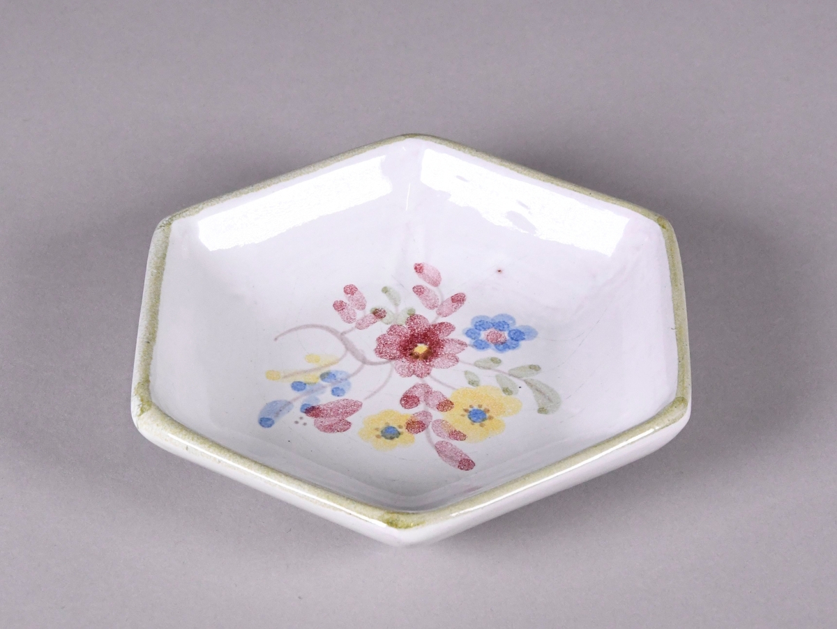 Skål av glassert keramikk, med opphøyd rand. Skålen er sekskantet, og har hvit grunnfarge. Håndmalt dekor av røde, gule og blå blomster på midten, og en grønn borde langs randen.