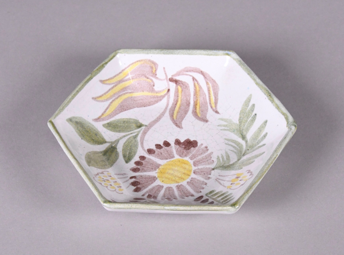 Skål av glassert keramikk, med opphøyd rand. Skålen er sekskantet, og har hvit grunnfarge. Håndmalt dekor av blomster og bladverk, lysebrun blomst med brune og gule blader, og en grønn borde langs randen.