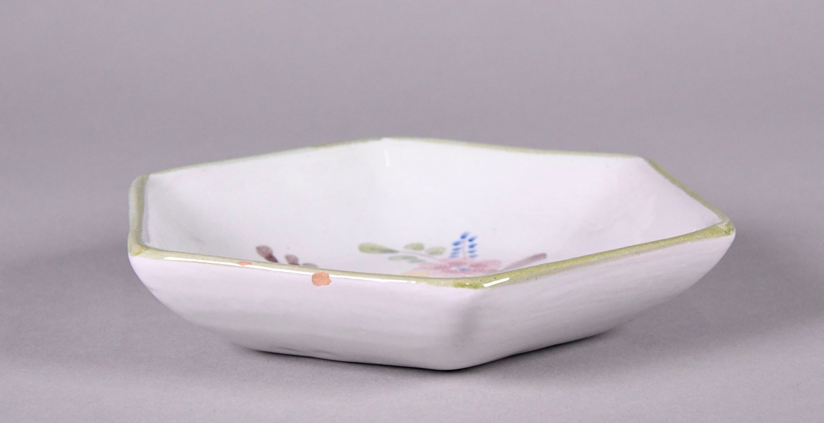 Skål av glassert keramikk, med opphøyd rand. Skålen er sekskantet, og har hvit grunnfarge. Håndmalt dekor av gul, rød og blå blomst, og en grønn borde langs randen.
