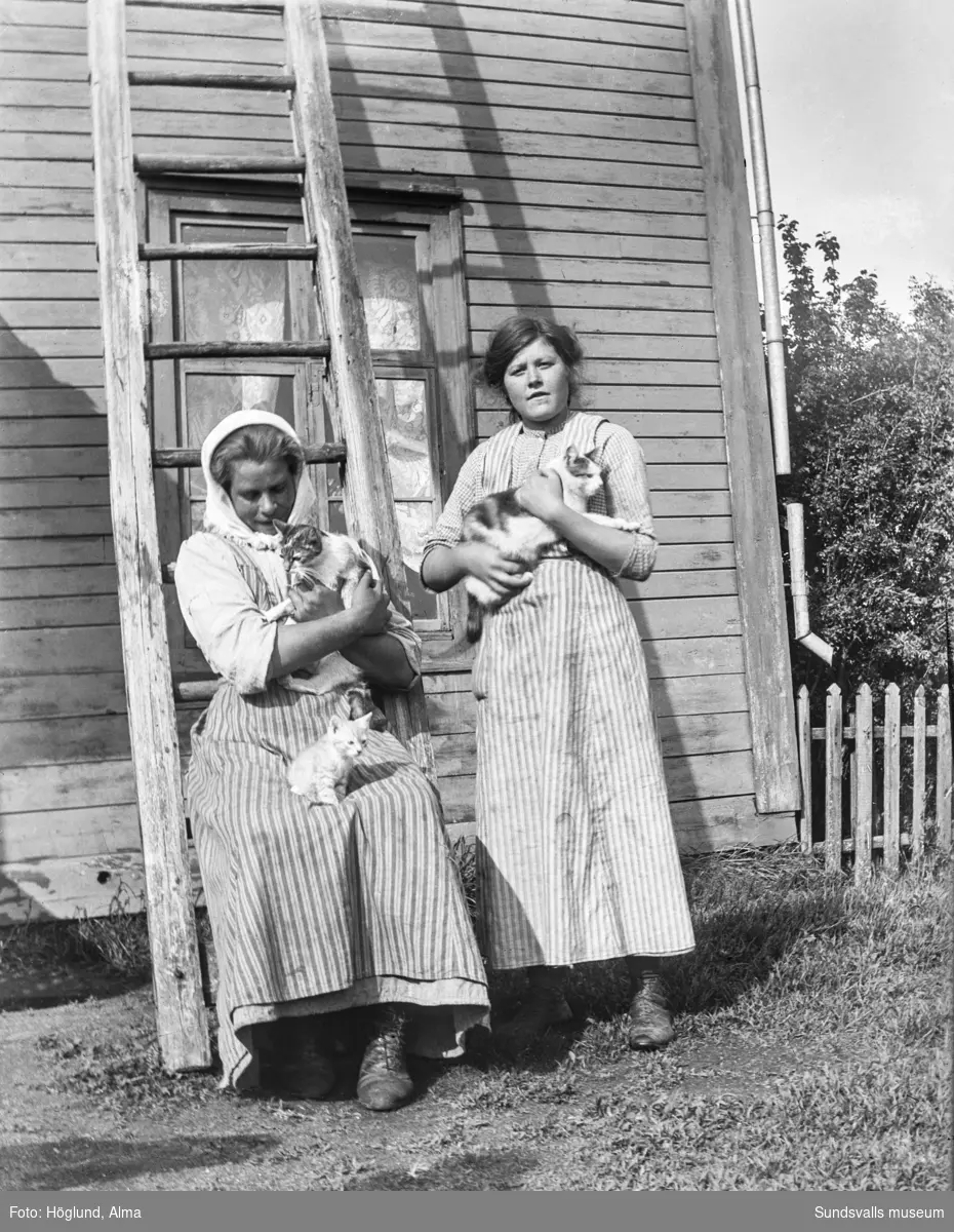 Två unga kvinnor med katter i famnen invid en stege som står lutad mot en husvägg.
