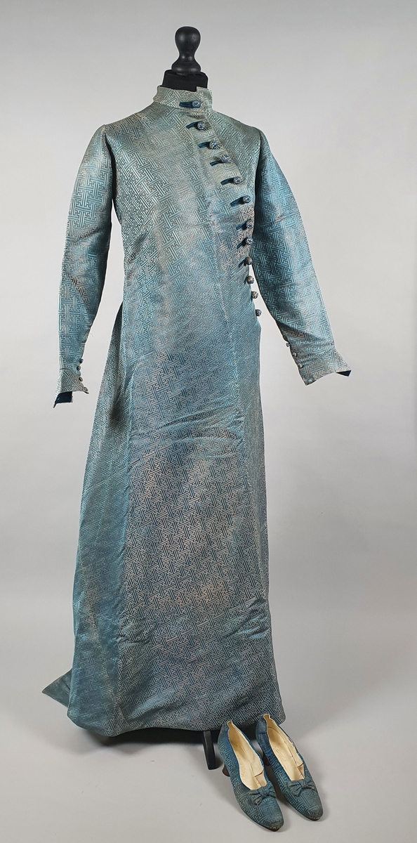 Turkis silkekjole med lange ermer og slep. Knapper diagonalt over brystet på venstre side. Föret med turkis silke.