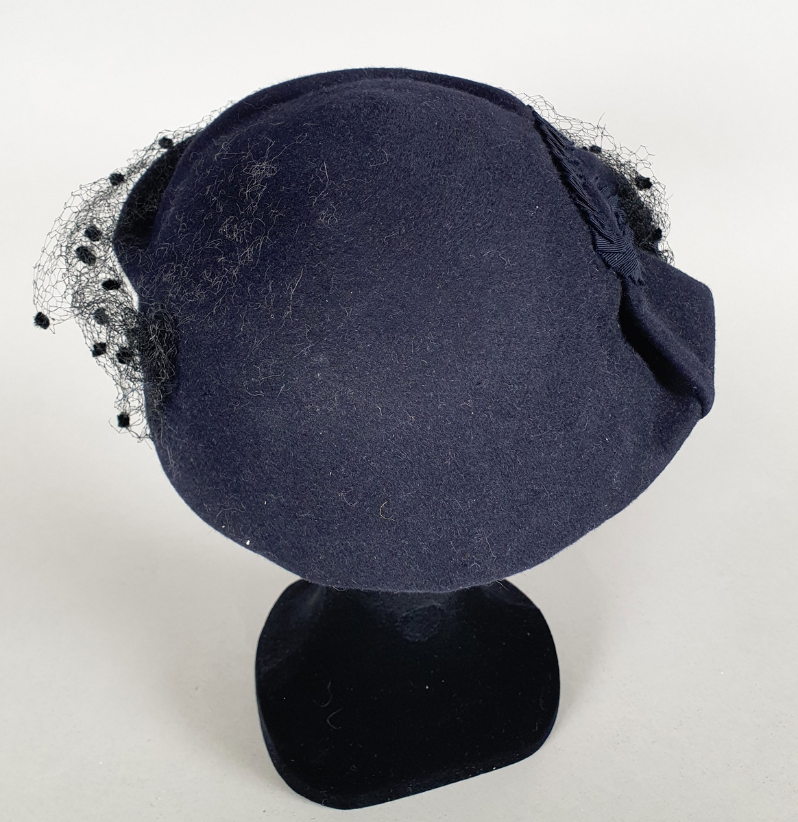 Mørkeblå damehatt med rund form og rosett på høyre side. Tynn netting med svarte ullprikker på forsiden.