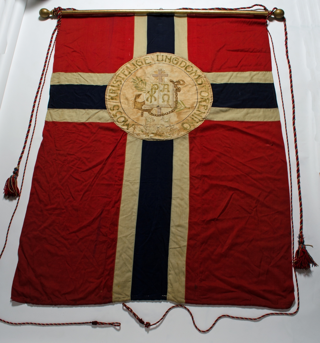 Faneduk i ull, norsk flagg i rødt, hvitt og blått med påsydd sirkel med tekst og dekor. På baksiden er det ikke påsydd sirkel med tekst og dekor. Faneduken er montert på tverrstang med kule i hver ende, to styresnorer og en bardunsnor.