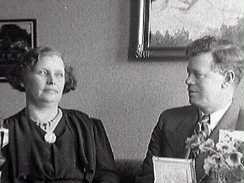 Födelsedagsfirande hos gelbgjutare Karl Holmkvist med fru. De sitter i soffan med blommor och presenter runtom.