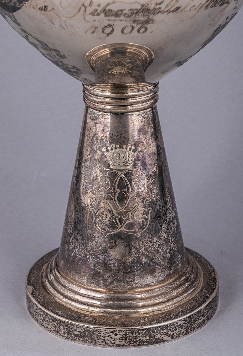 Pokal, silver, inskription: HKH Kronprinsens Hederspris vid Svenska Riksskyttetävlingen 1906. 
På foten kronprinsens krönta spegelmonogram.