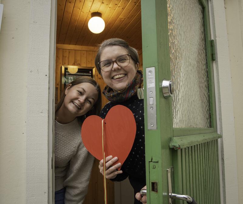 To jenter står i døra og ønsker velkommen inn. Den ene holder et hjerte i hånden.