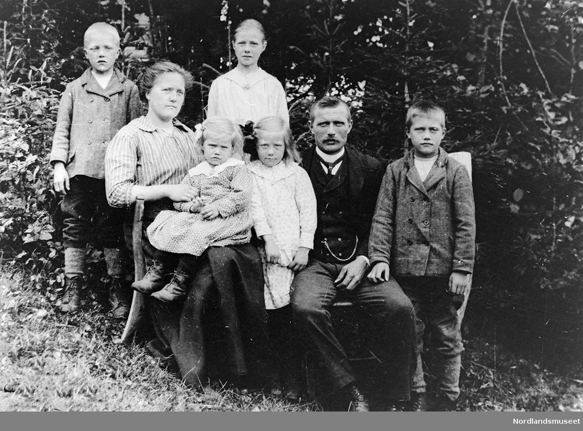 Albert og Amalie Rønnebekk med familie. De sitter i en skråning, barna står bortsett fra ett barn som sitter på fanget til Amalie. Tett buskas bak dem.