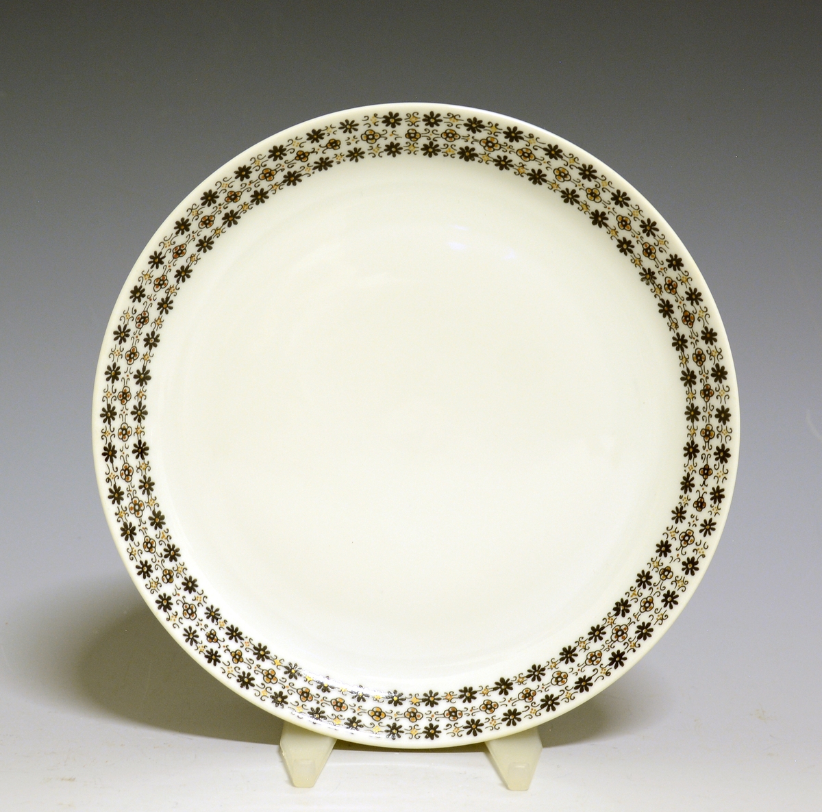 Asjett av porselen med hvit glasur og bord med småblomstret trykkdekor i sort og gull langs kanten.
Modell: Petita (eller variant av)

