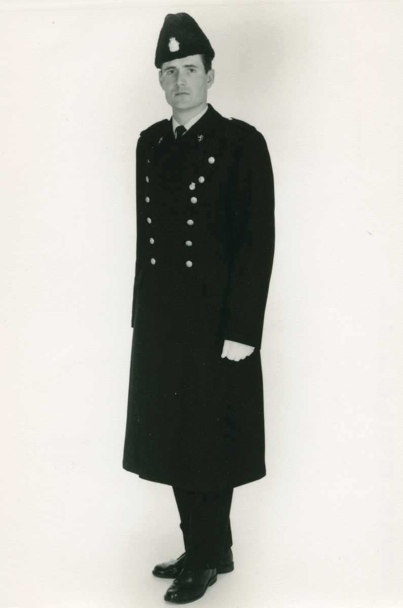 Politimann iført politiuniform, vinterlue M/1935 og helårskappe. Kappen fra 1953-reglementet og 1963-reglementet har samme snitt.