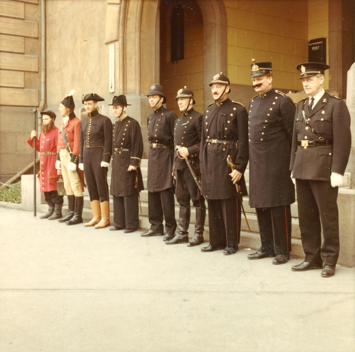 Ni menn utkledd i historiske politiuniformer fra Oslo, fra 1700-tallets vekteruniform til politiuniform modell 1964. Mennene er oppstilt utenfor hovedinngangen til Oslo hovedpolitistasjon i Møllergata 19.