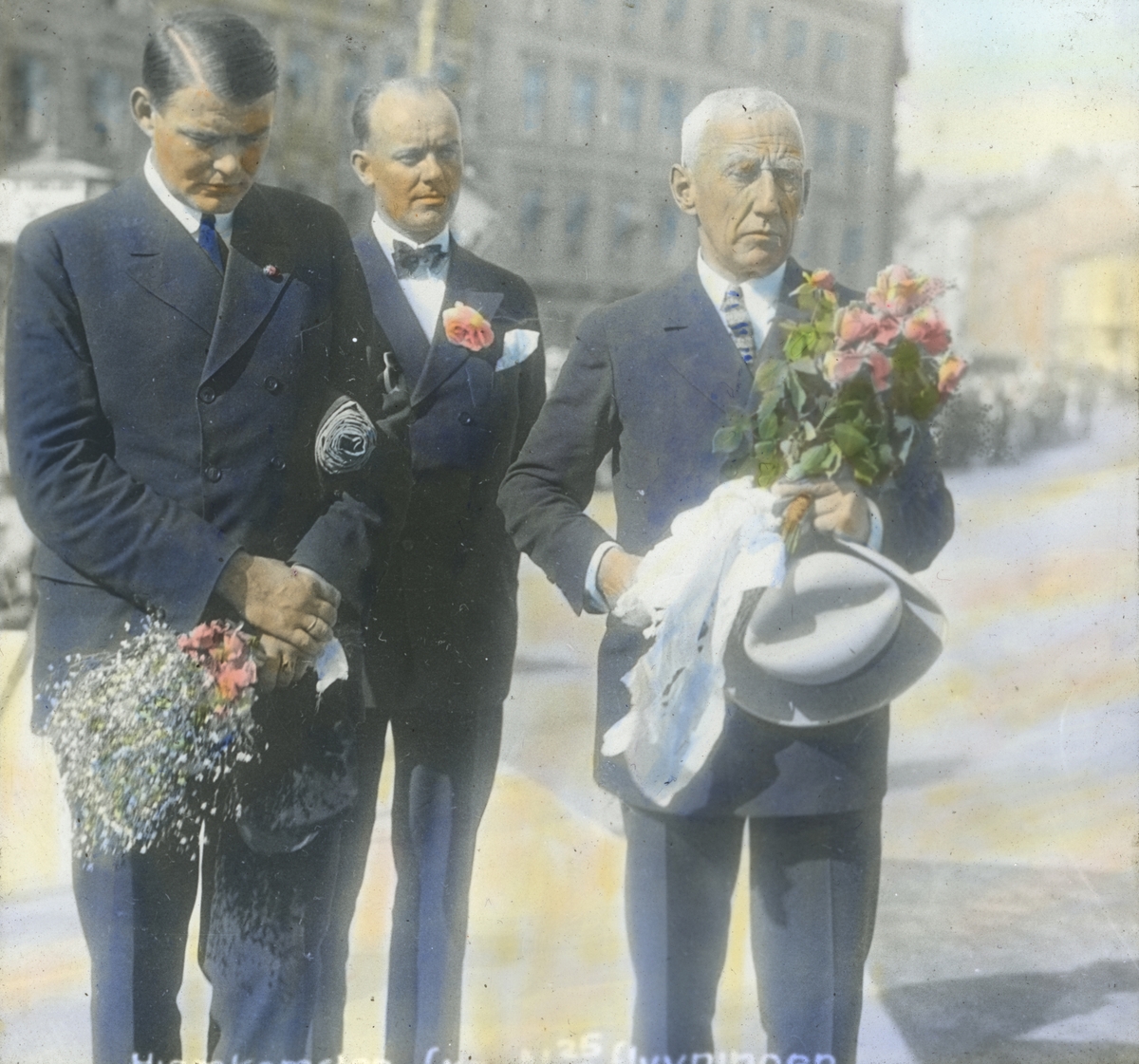 Håndkolorert dias. Mannskapet på de to flyene "N24" og "N25" ble feiret som helter under mottakelsen i hovedstaden. Roald Amundsen og Hjalmar Riiser-Larsen har blomster i hendene. En mann står bak dem. 