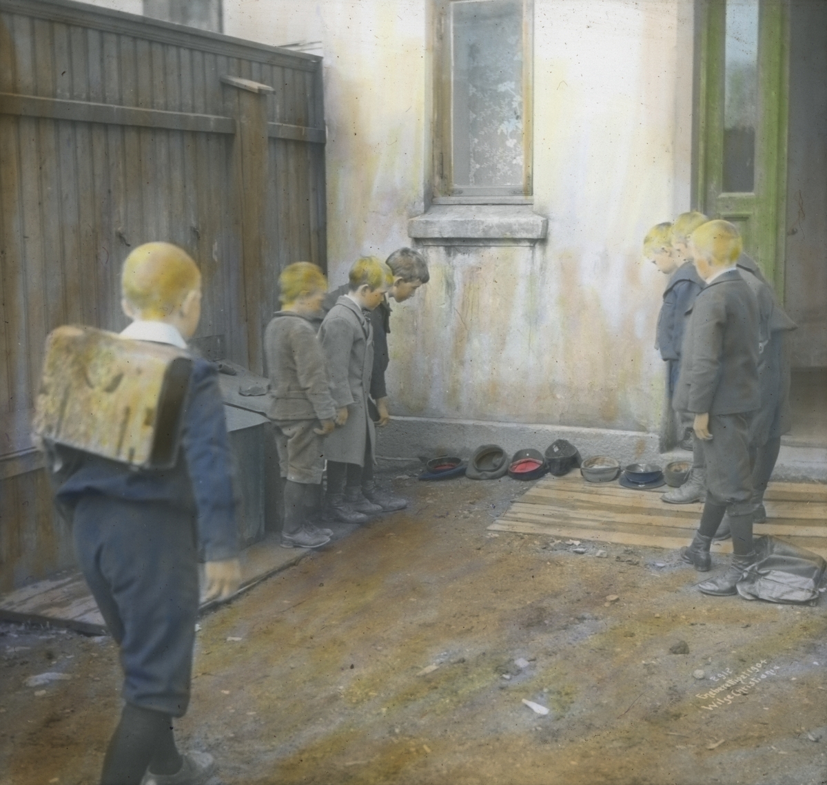 Håndkolorert dias. 7 gutter leker i en bakgård i 1904. Luene deres ligger på rekke ved veggen, 6 av guttene ser på disse, mens en gutt med ransel står klar til å kaste.