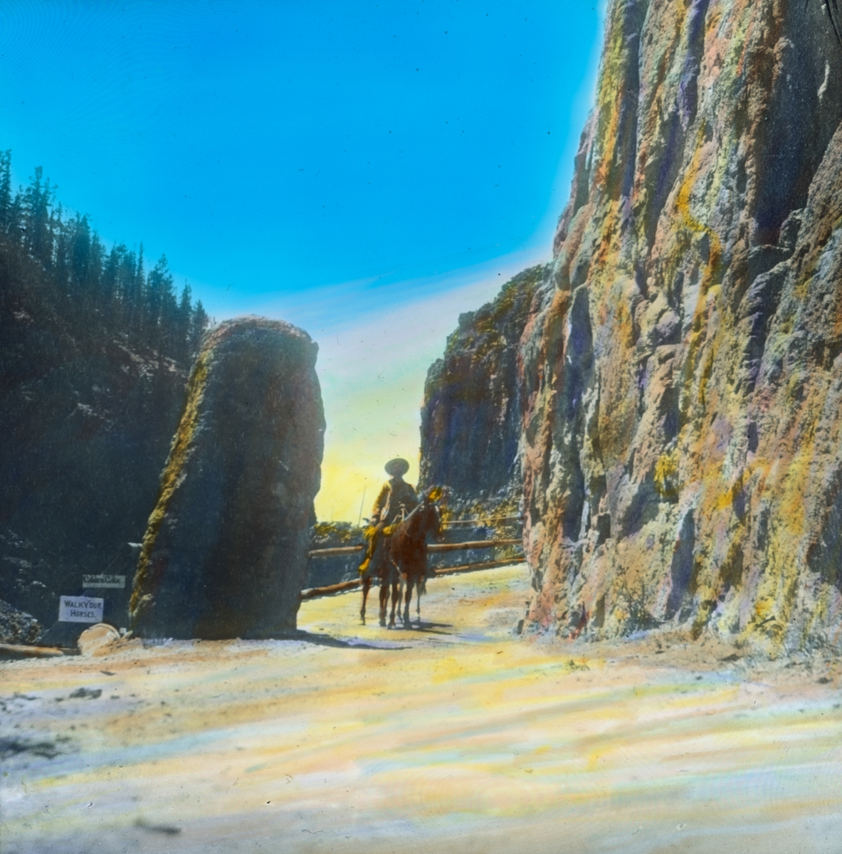 Håndkolorert dias. En mann sitter i sadlen på en hest, som står på en grusvei mellom en bratt fjellside og en høyreist stein i dalen Golden Gate, Yellowstone nasjonalpark. To skilt står ved steinen med påskriftene "Golden Gate" og "Walk your horses".  