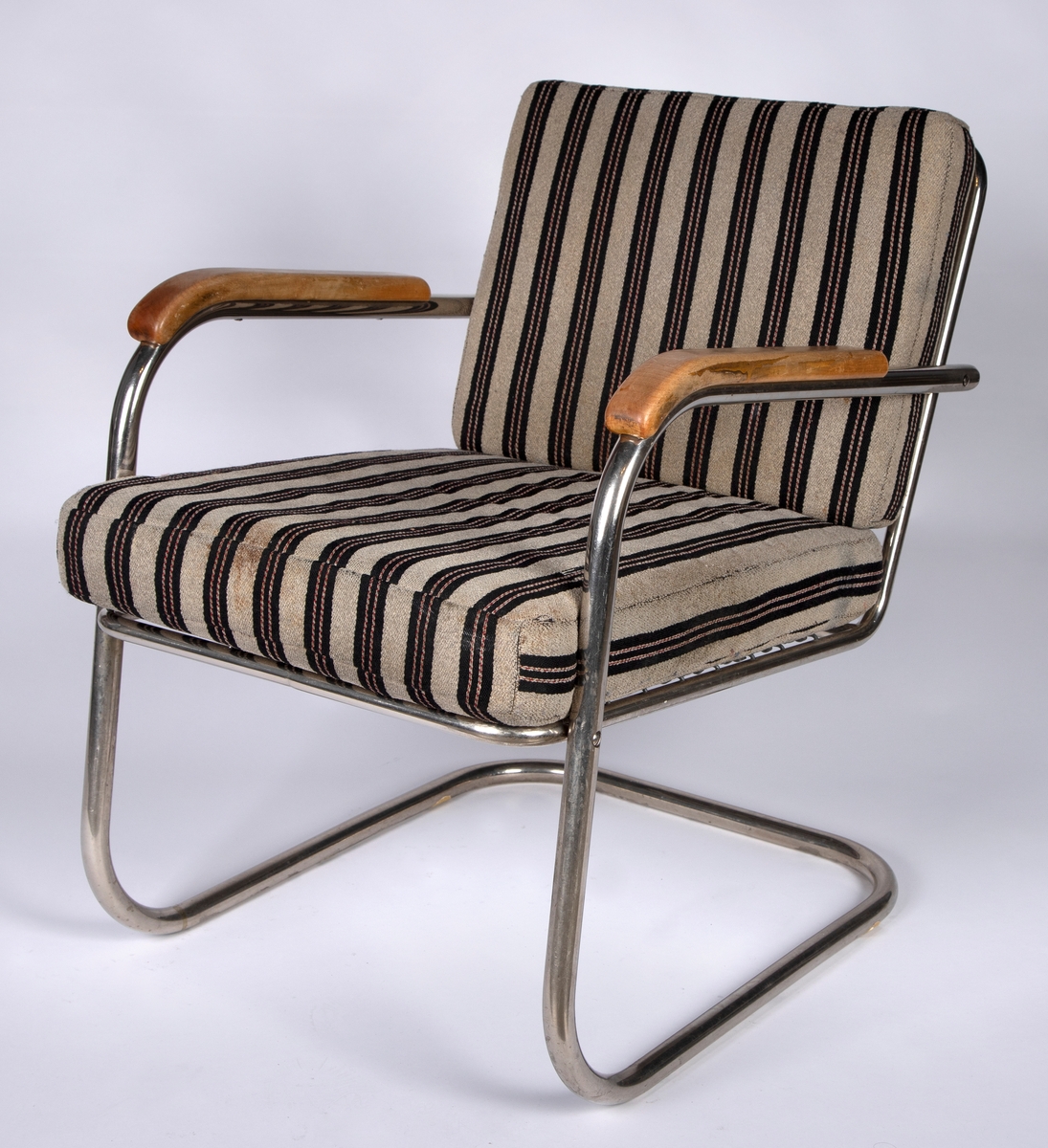 Stålrørslenestol med armlener i bjørk. Stolen er satt sammen av to stålrørselementer. Ben og lener er ett element og sete og rygg er ett element. Festepunkt midt på ryggen og foran på setet. Tre stålrørssprosser i ryggen og fjæring i sete. Gråbrunt møbeltrekk med vertikale brede, svarte striper som har to parallelle rader med smale vertikale striper. Disse er gråbrune med stiplete røde striper.