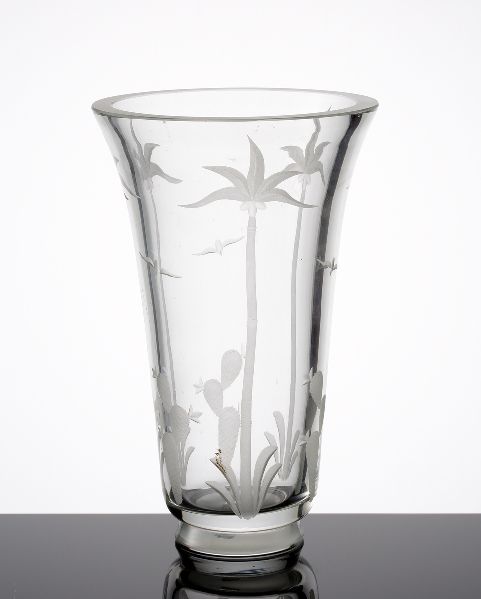 Formgiven av Edward Hald. Hög vas i tjockt glas med klack. Mattslipad dekor på kupan föreställande palmer,
kaktusar och fåglar.