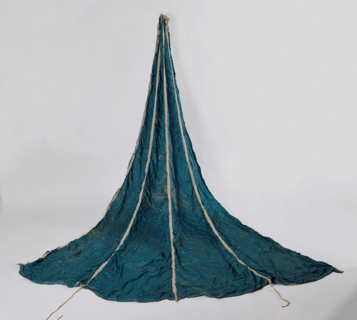 Del av fallskjerm i blå-turkis silke. Trekant forma, sydd saman av fire delar. Merka med 1 2 3 4 ved kvar del. Påsydde kvite band over kvar skjøyt. Kvite snorer