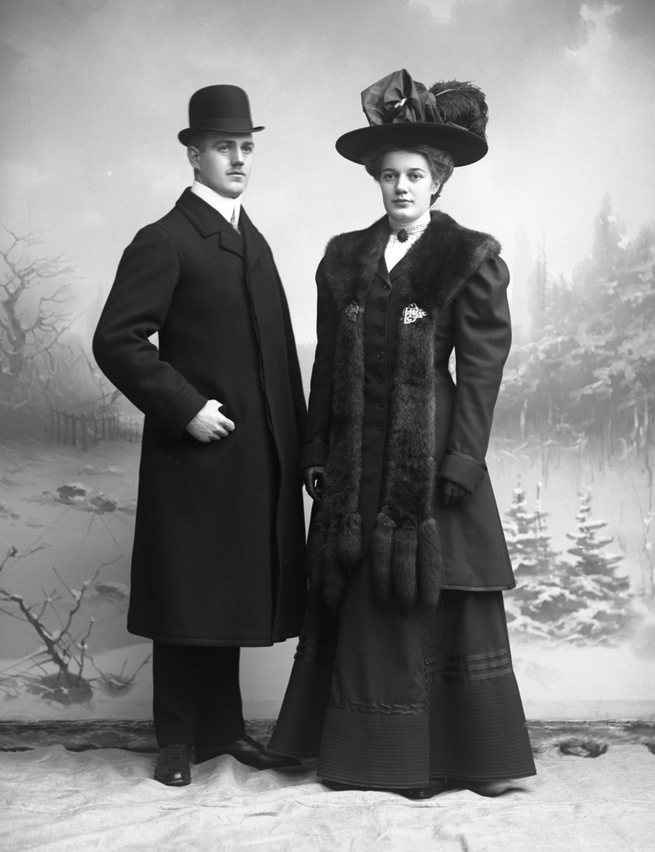 Ateljébild av en man och en kvinna i vinterkläder mot fond med snöigt landskap. Kvinnan bär en bred pälskrage med spännen framtill och långa, fritt hängande ändar kantade med tre svansar vardera.