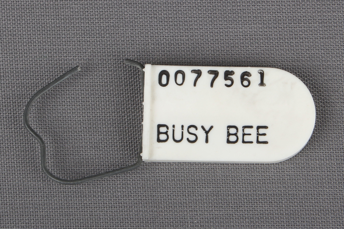 Hvit plastbrikke med svart tekst. En ståltråd er sitter fast i merket (trolig til å feste på bagasje), og senere klipt av.