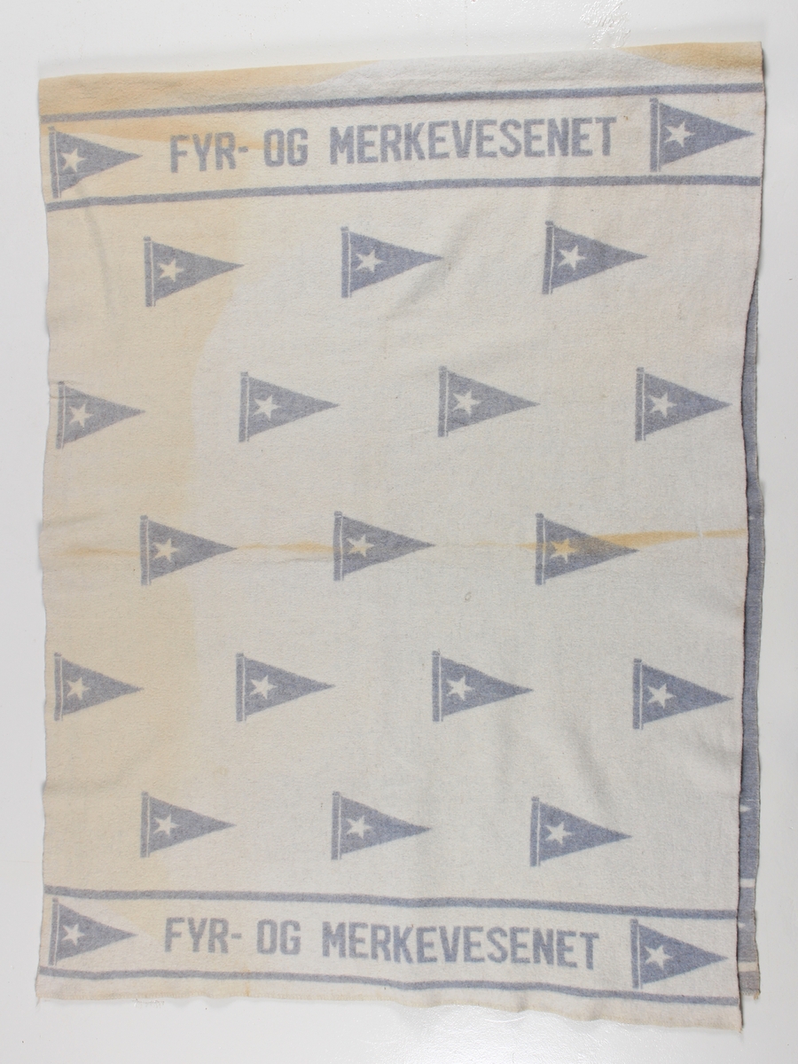 Hvitt og lyseblått ullteppe med vevd motiv som viser logoen til Fyr- og merkevesenet: Et trekantet flagg med femkantet stjerne.
