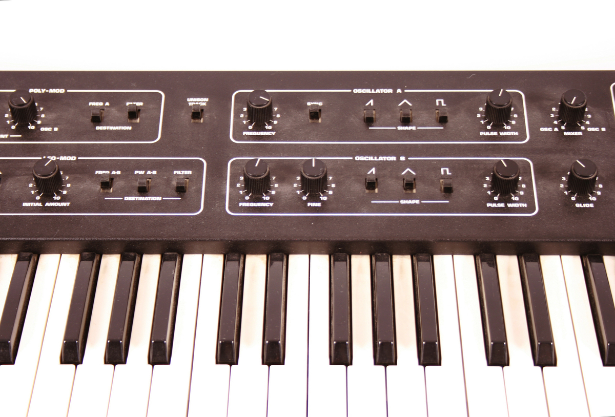 Elektronisk musikkinstrument, synthesizer. Tastatur med klaver nederst mot brukeren. Øverste rekke består av vridbare knapper. På vesntre side et panel med tall. Baksiden har inngang til ulike ledninger.