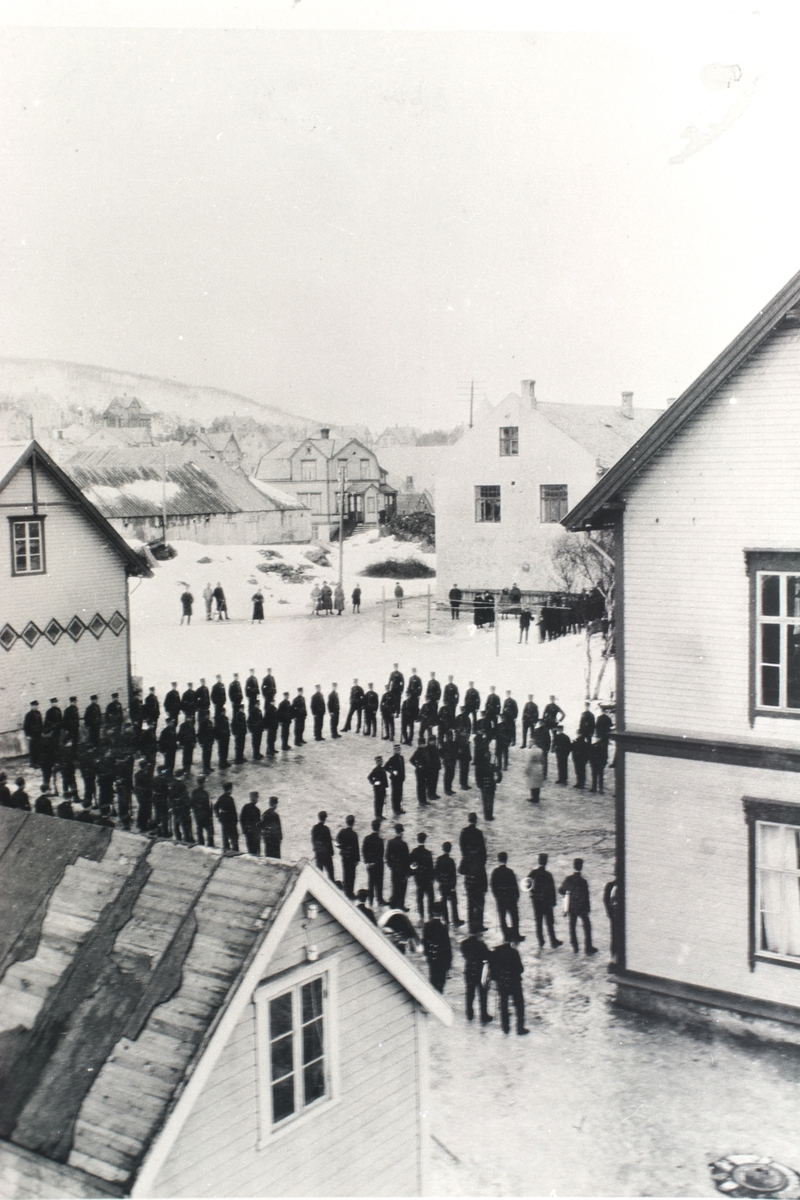 Soldater oppstilt på en plass mellom flere bygninger.