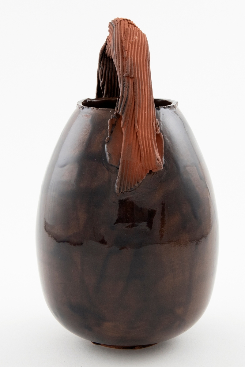 Blank brunglasert vase i terrakotta med eggformet korpus. Lav fot og åpen munning. Vasen har en skulpturell hank i matt brun terrakotta med riller. Hanken går i en bue over munningen.