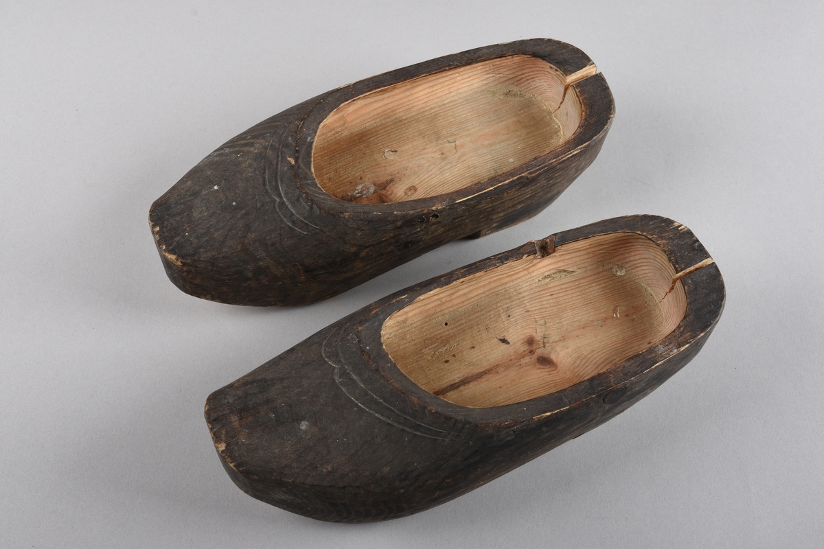 Tilskore sko av tre. A) er venstre sko og B) er høgre sko. Begge er tjærebehandla på utsida, men ubehandla inni og under sålen. Dei er brukt og ein del nedslite under sålen. A har ein kvist heilt oppe i kanten på høgre side og eit hakk i botn like nedanfor kvisten. Dessutan er det ein kvist under hælen. B har to kvistar i sålen rett foran hælen. Begge er sprukke i overkant av hækappa og har hol i sida til å buntast saman med.