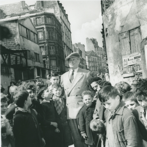 Maurice Chevalier og en flokk med unger. I bakgrunnen bygninger. 