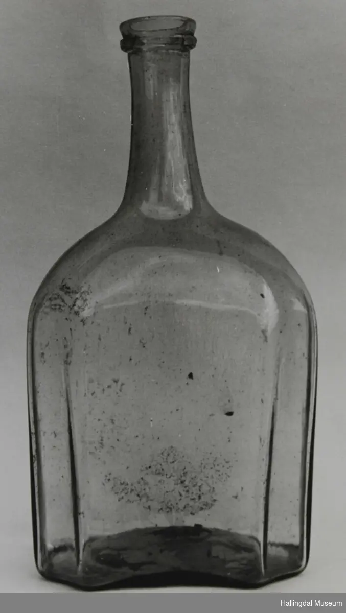 Flaske i ufarga glas. 8-kanta botn, 8-kanta tverrsnitt, oval opp til halsen som har sirkulært tverrsnitt og er smalast på midten.