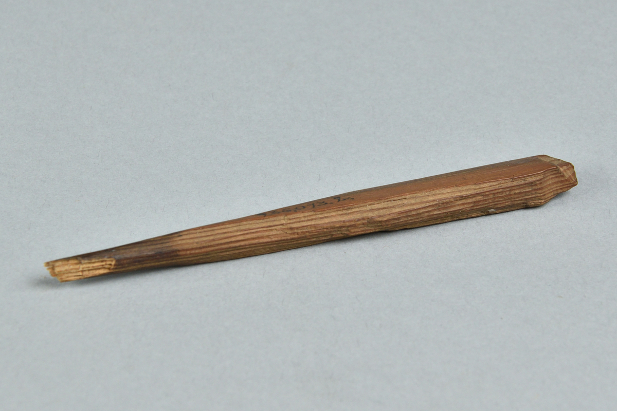 Fragment av sticka tillverkad i trä, täljd. Endast det platta avslutet av stickan samt en kort bit av skaftet finns kvar.