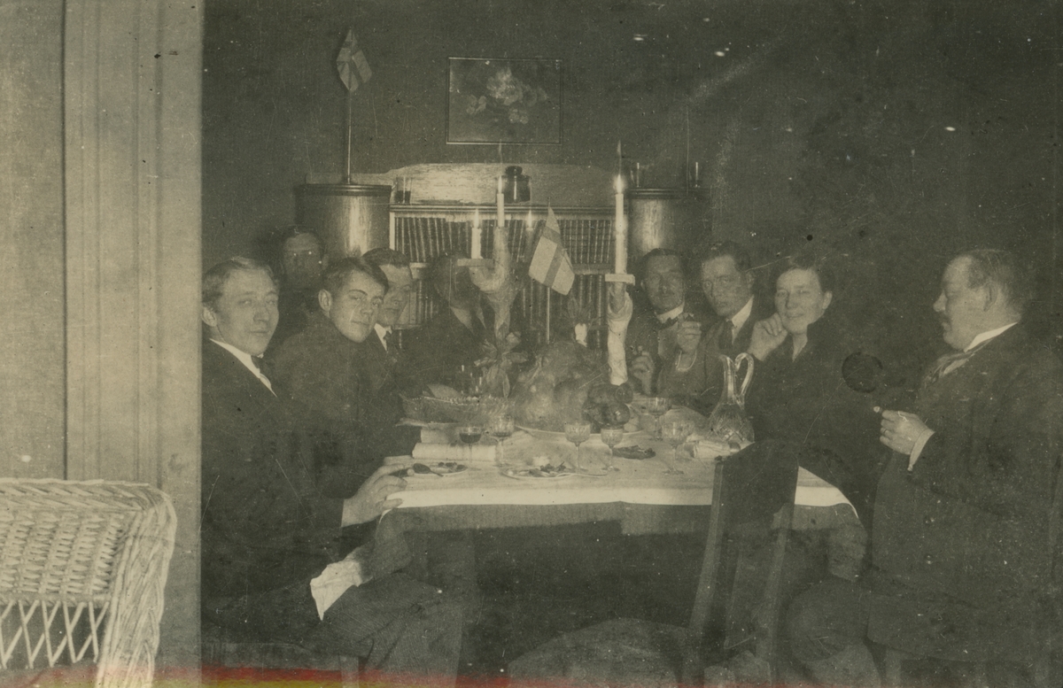 Fotografi från expedition till Spetsbergen. Motiv av åtta finklädda män och en kvinna som sitter runt ett bord och äter middag. På bordet finns ett stort grishuvud med äpple i munnen och flera vinglas, ljusstakar och en flagga.