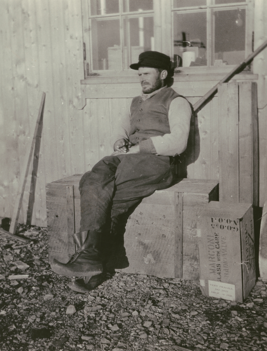 Fotografi från Ahlmannexpeditionen 1931. Motiv av man som sitter och vilar på trälåda framför hus.