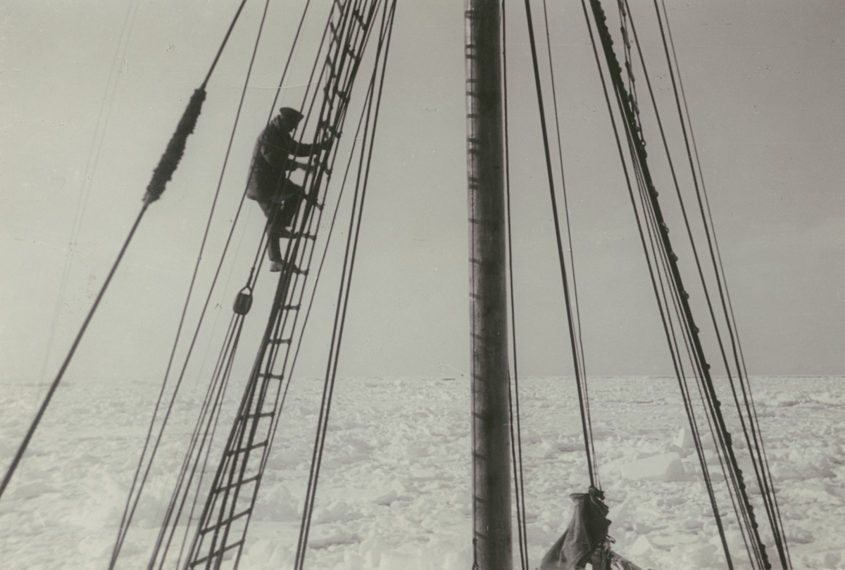Fotografi från svenska undsättningsexpeditionen 1928. Motiv av fartyg med man som klättrar nerför stege. Fartyget är omgivet av ett islandskap.
