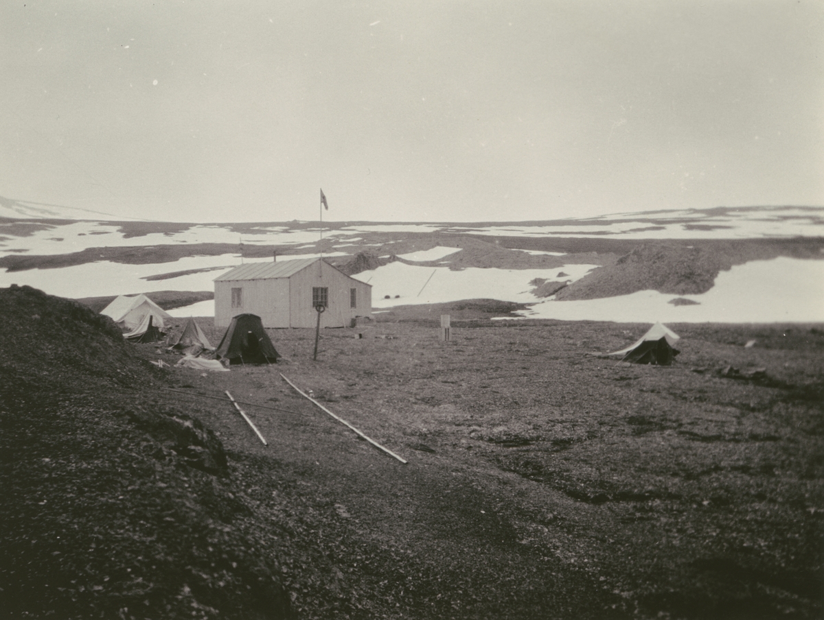 Fotografi från Ahlmannexpeditionen 1931. Motif of a small house and tents in a snow covered landscape.
