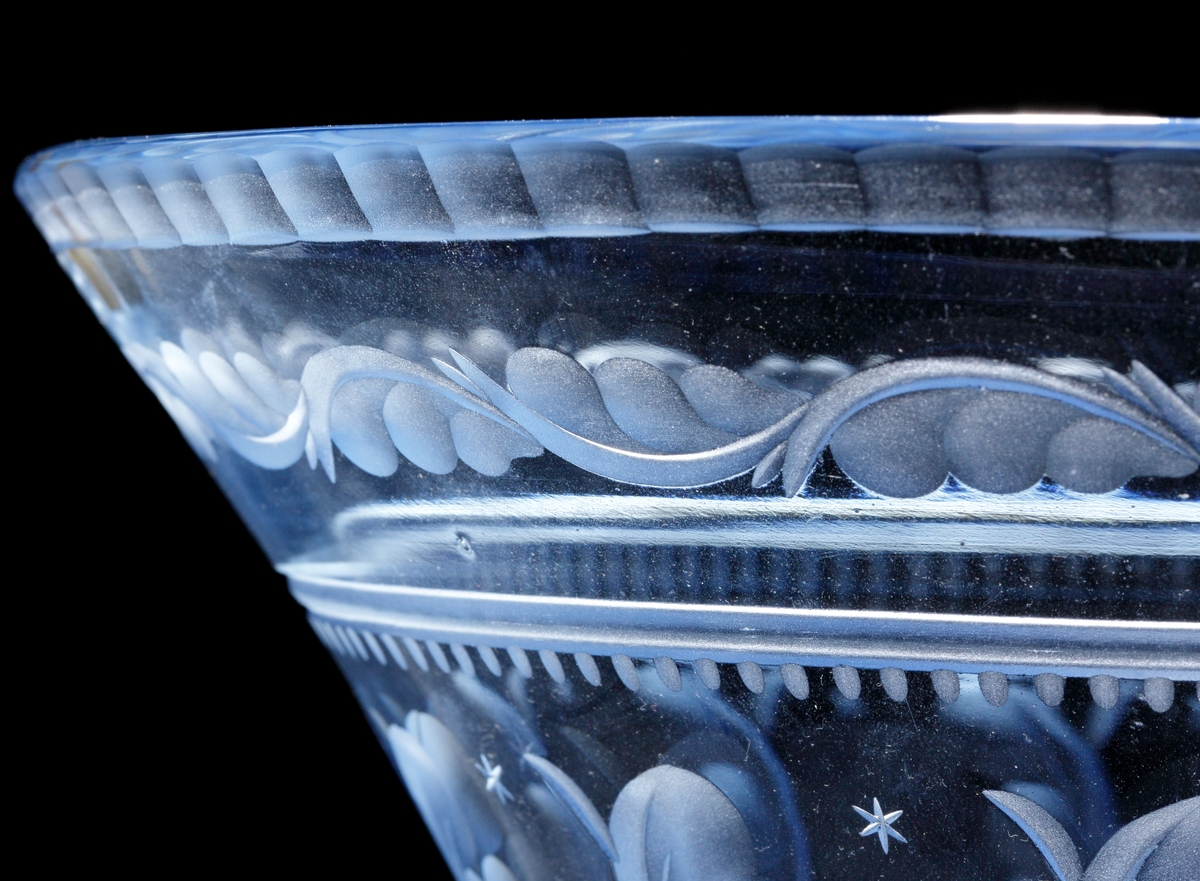 Formgiven av Edward Hald. Ljusblå trattformad skål med fasetterad mynningskant, slipad fjäderformad dekor och
små stjärnor.