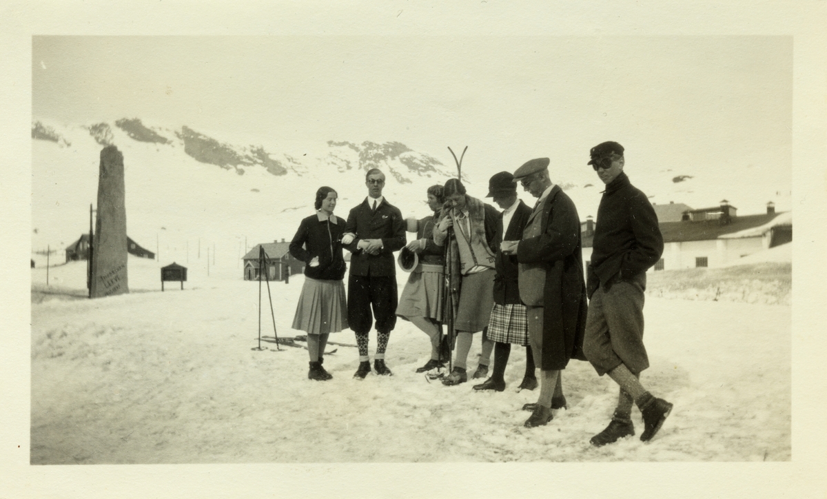 En gruppe påsketurister fotografert før avreisen fra Finse. Blant dem er antagelig Lucy Egeberg og Anton Fredrik Klaveness. Til venstre obelisk til minne om polfareren Robert Scott. Fotografert påsken 1928.