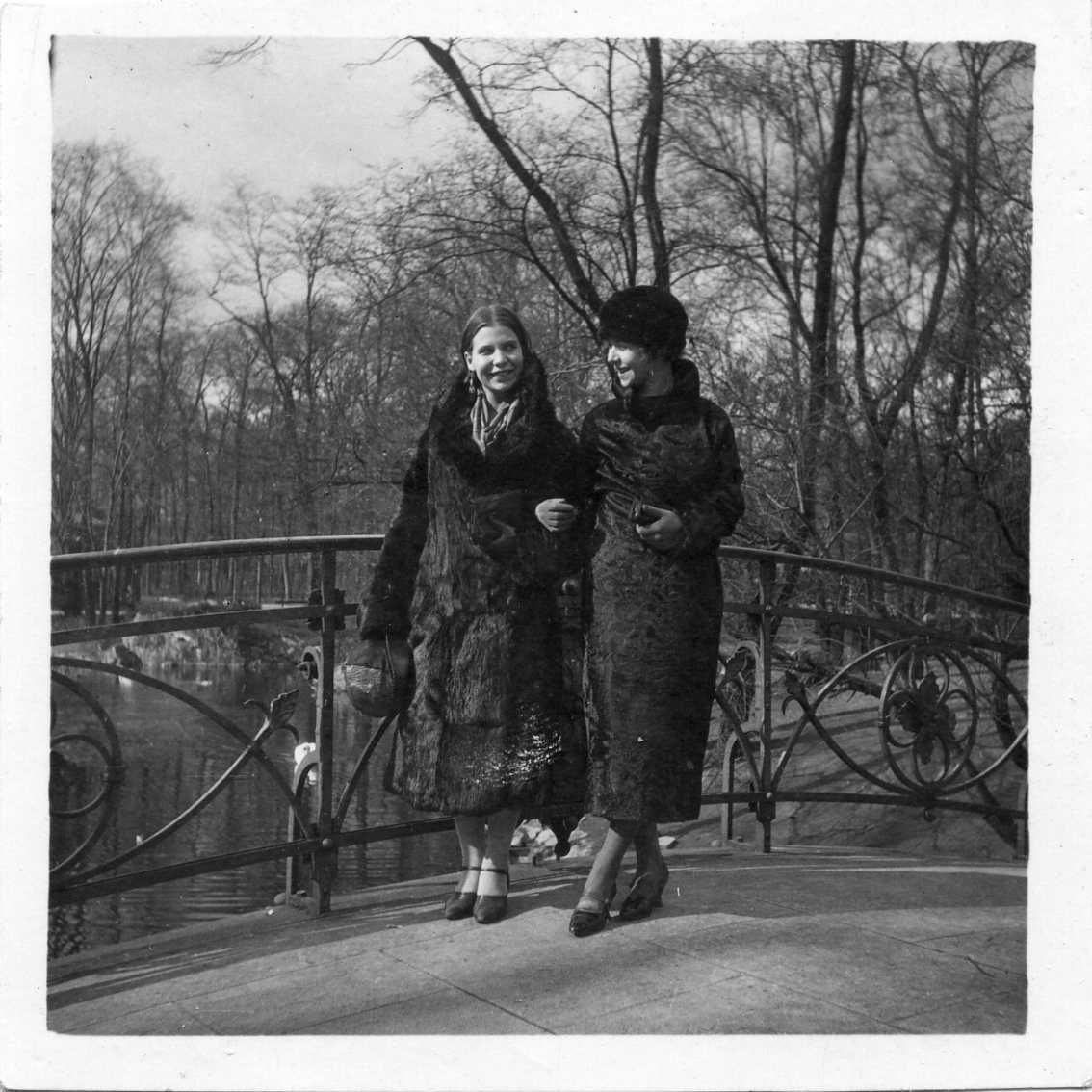 Två kvinnor i mörka kappor står armkrok på en bro med järnräcke i jugendstil. Det är vårvinter.