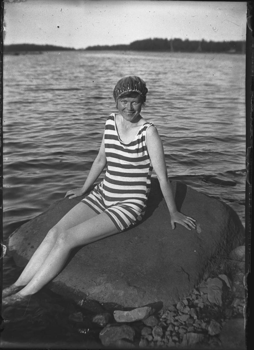 En kvinna i randig baddräkt, på en sten i sjön.