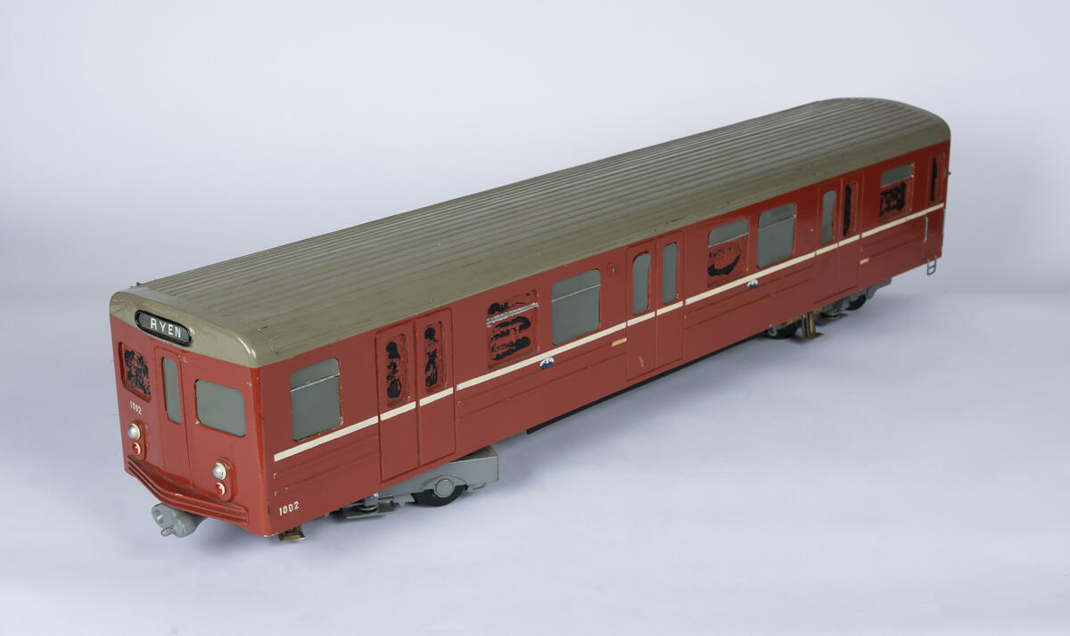 Tremodell av T-banevogn nr. 1002 i en rød farge, hvit stripe langs sidene og grått tak. Under er modellen uthulet og og det er plassert to "boggier" i hver ende. 