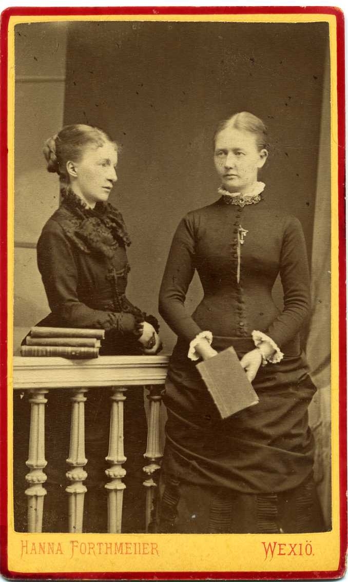 Två unga kvinnor står vid ett räcke i en ateljé. Det ligger böcker på räcket och kvinnan till höger håller en bok i händerna.