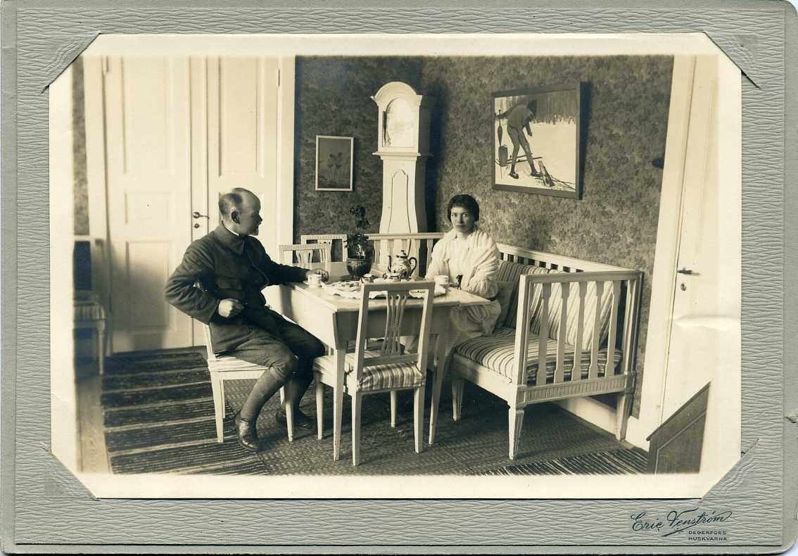 En kvinna och en man sitter och dricker kaffe i sitt hem. Mannen sitter på en stol och kvinnan sitter i en soffa mittemot. På väggen ovanför henne hänger en målning av mannen.