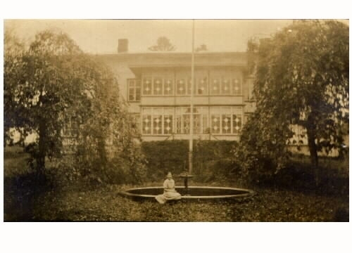 Mangårdsbyggnaden på Torstorps gård i Grimeton. Byggnaden har en står glasveranda i två våningar. Mitt framför den står en flaggstång vid en rund fontän, där en ung kvinna sitter på kanten.