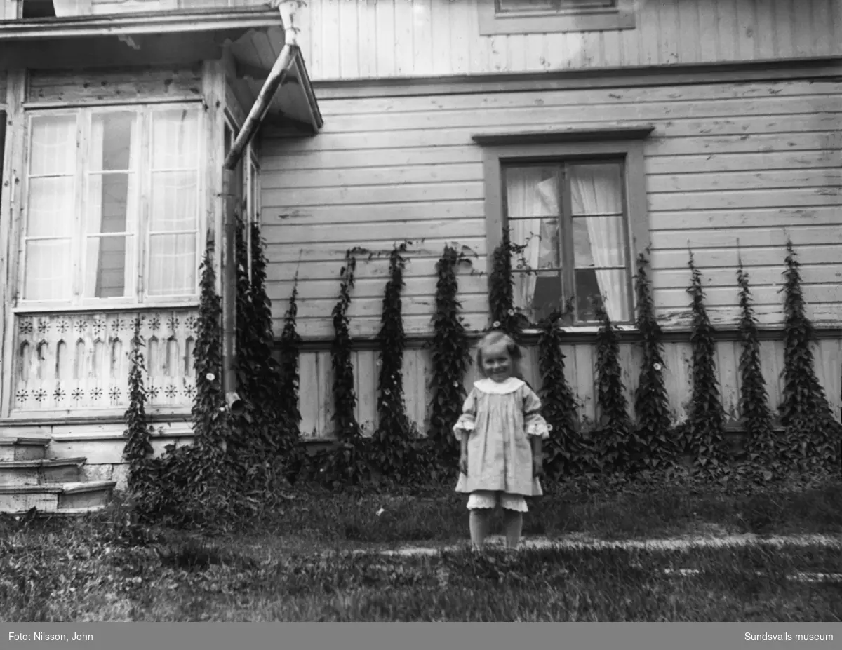En liten flicka står invid glasverandan på Roken 1:7 i Laggarberg. En rad med klätterväxter pryder husväggen.
