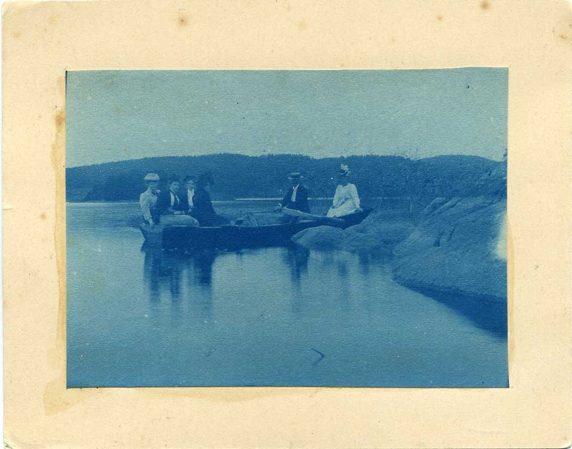 Fem kvinnor och en man i en roddbåt vid en klippstrand.